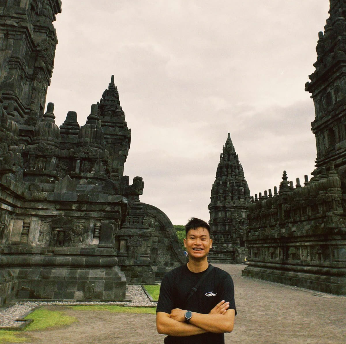Clausewitz standing with his arms folded on the stone avenue between the temples at Candi Prambanan, under an overcast sky.